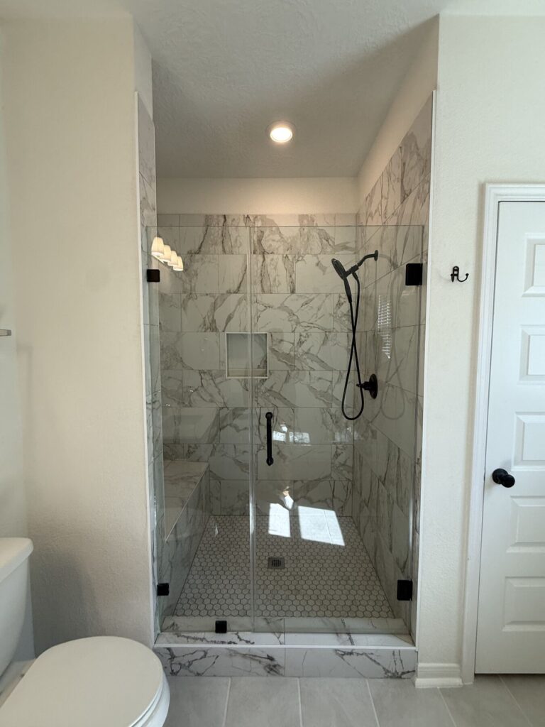 Glass-enclosed shower with white marble tiles, pebble floor, and black fixtures including a handheld showerhead; toilet and entry door visible nearby.