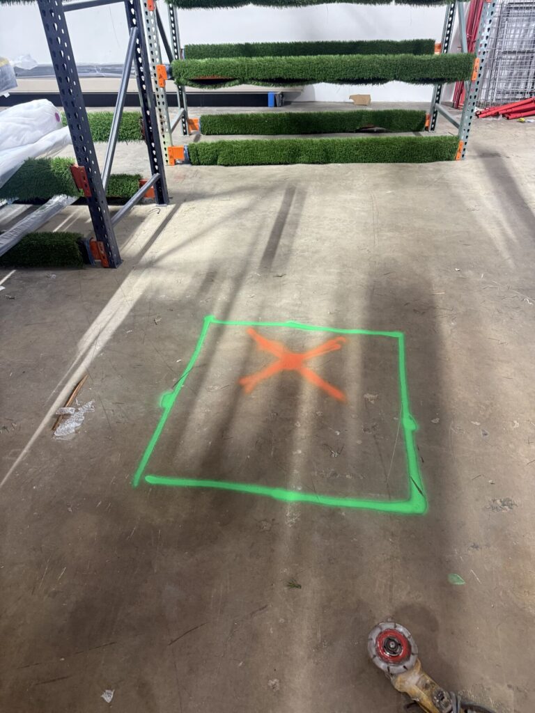 Warehouse with turf rolls on metal racks; a neon green square with an orange X marked on the floor indicating a restricted area.